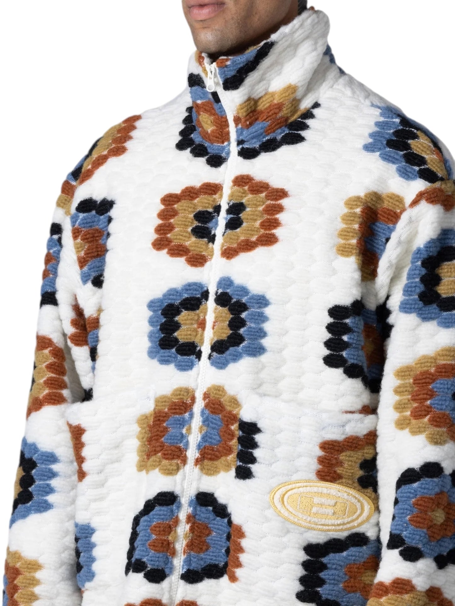 A person is wearing the HONOR THE GIFT TEXTURED FULL ZIP CREAM jacket, featuring a colorful abstract pattern with blue, brown, and orange accents. The jacket boasts a novelty texture fleece design and an emblem on the lower left front, giving it a festival-inspired vibe.