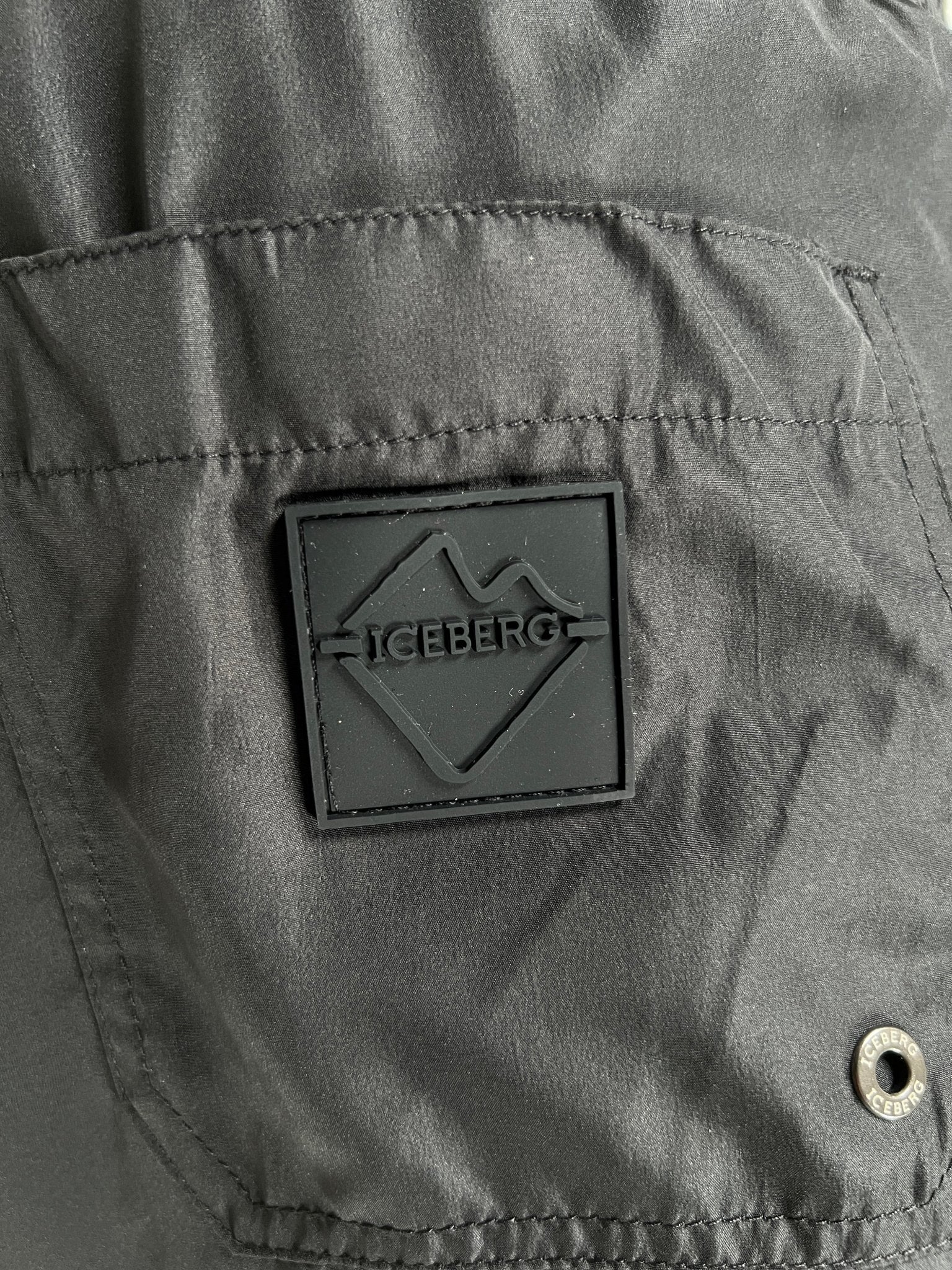 Close-up of the Iceberg ICE25MBM01 Medium Boardshort in black by ICEBERG, featuring a square patch with a stylized mountain and "ICEBERG" logo, plus a metal snap button detail on the lower right.