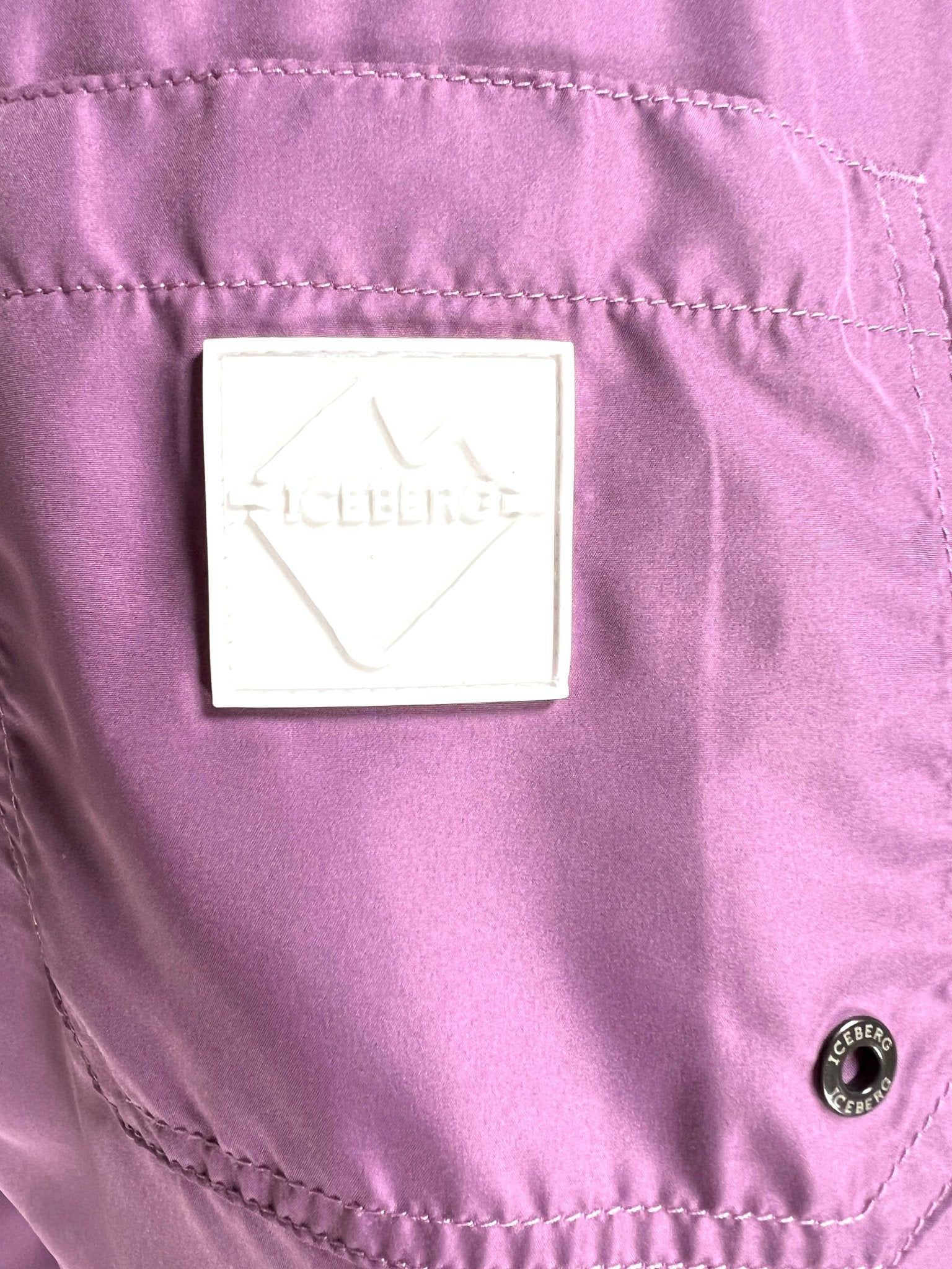 Close-up of a raspberry polyester ICEBERG ICE25MBM01 Medium Boardshort, featuring a white rectangular "ICEBERG" patch and a small round metal "ICEBERG" button.