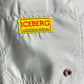 Close-up of the Iceberg ICE25MBS05 Short Boardshort Mint by ICEBERG, showing a light gray pocket in imported 100% polyester with a yellow and red "ICEBERG" patch above a metal button. Patch reads: "THE SUCCESS ABOVE & THE PERSISTENCE.