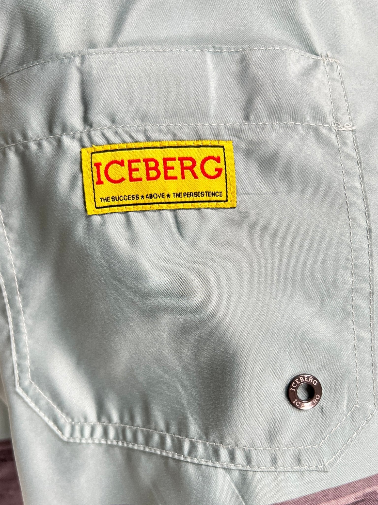 Close-up of the Iceberg ICE25MBS05 Short Boardshort Mint by ICEBERG, showing a light gray pocket in imported 100% polyester with a yellow and red "ICEBERG" patch above a metal button. Patch reads: "THE SUCCESS ABOVE & THE PERSISTENCE.