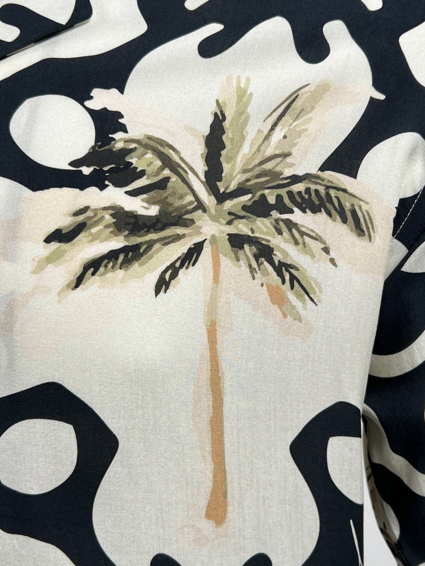 Close-up of a fabric with a black and white abstract pattern and a central print of a palm tree, featuring green fronds and a brown trunk, exuding a tropical vibe. Part of the INIMIGO collection's INIMIGO ISH3525 PALM SHIRT BLACK.