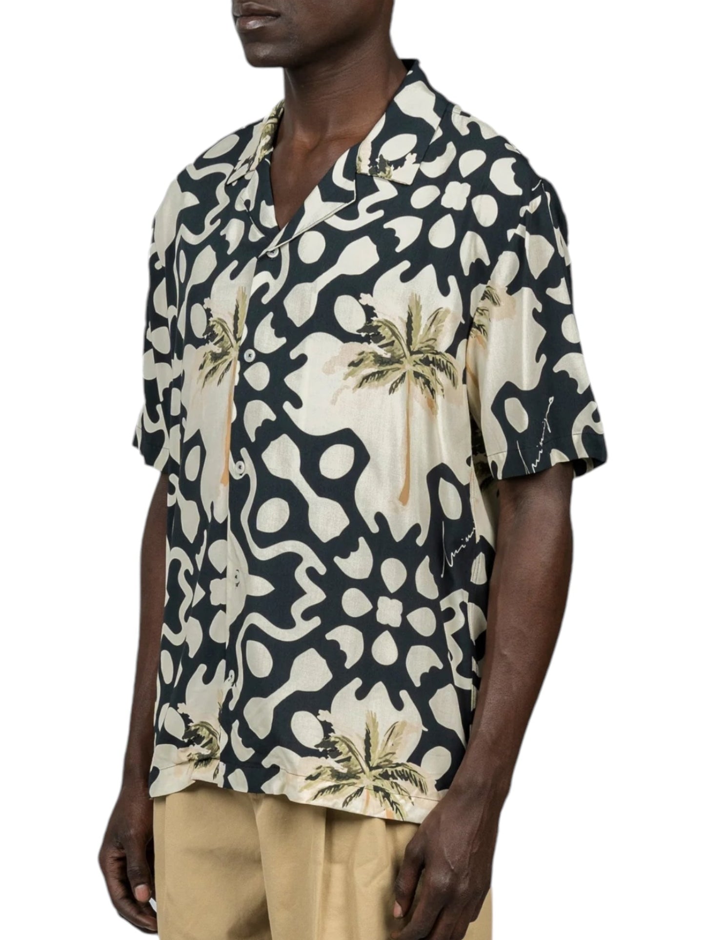 A person wearing the Inimigo ISH3525 Palm Shirt in black from the INIMIGO collection, featuring a short-sleeve design with a cream abstract pattern and palm tree motifs, paired with light brown pants.