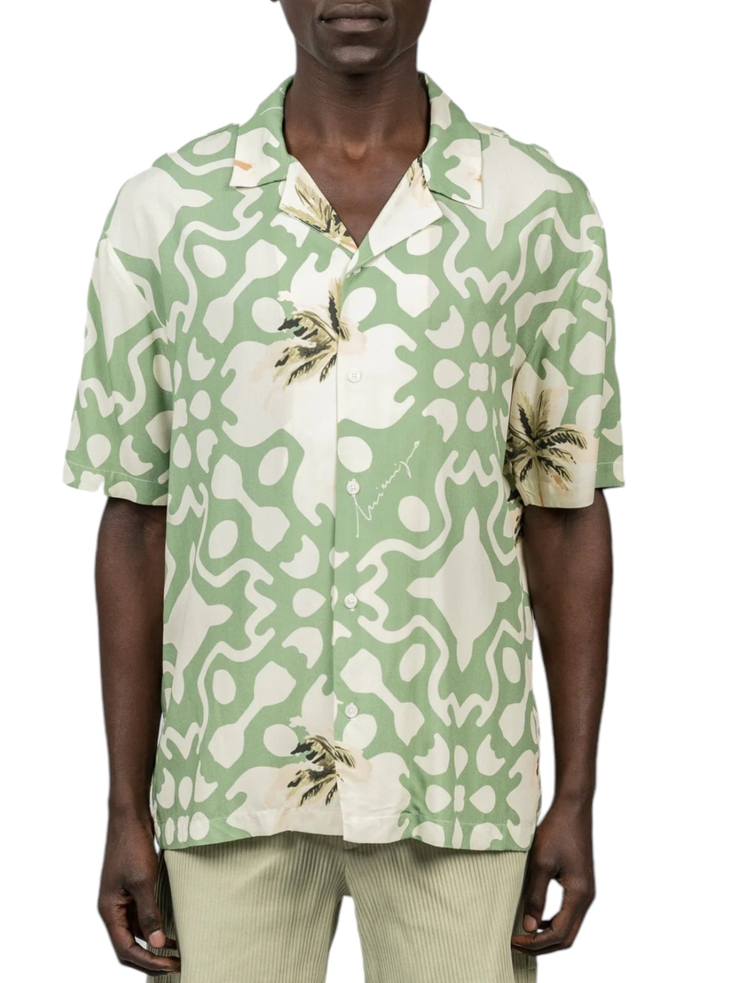 The individual is wearing the INIMIGO ISH3525 Palm Shirt Green, featuring abstract palm tree designs in a striking green and white pattern, complemented by beige pants. This versatile button-up shirt from INIMIGO adds a fresh vibe to the ensemble, making it perfect for both casual outings and relaxed summer gatherings.