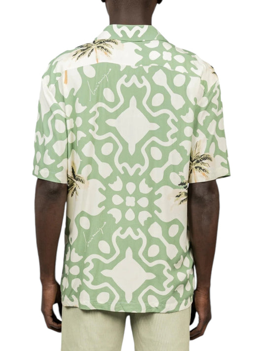 A person wears the Inimigo ISH3525 Palm Shirt Green by INIMIGO, a short-sleeve tropical shirt showcasing a green and white abstract pattern with a palm tree design on the back, perfectly matched with light-colored pants.