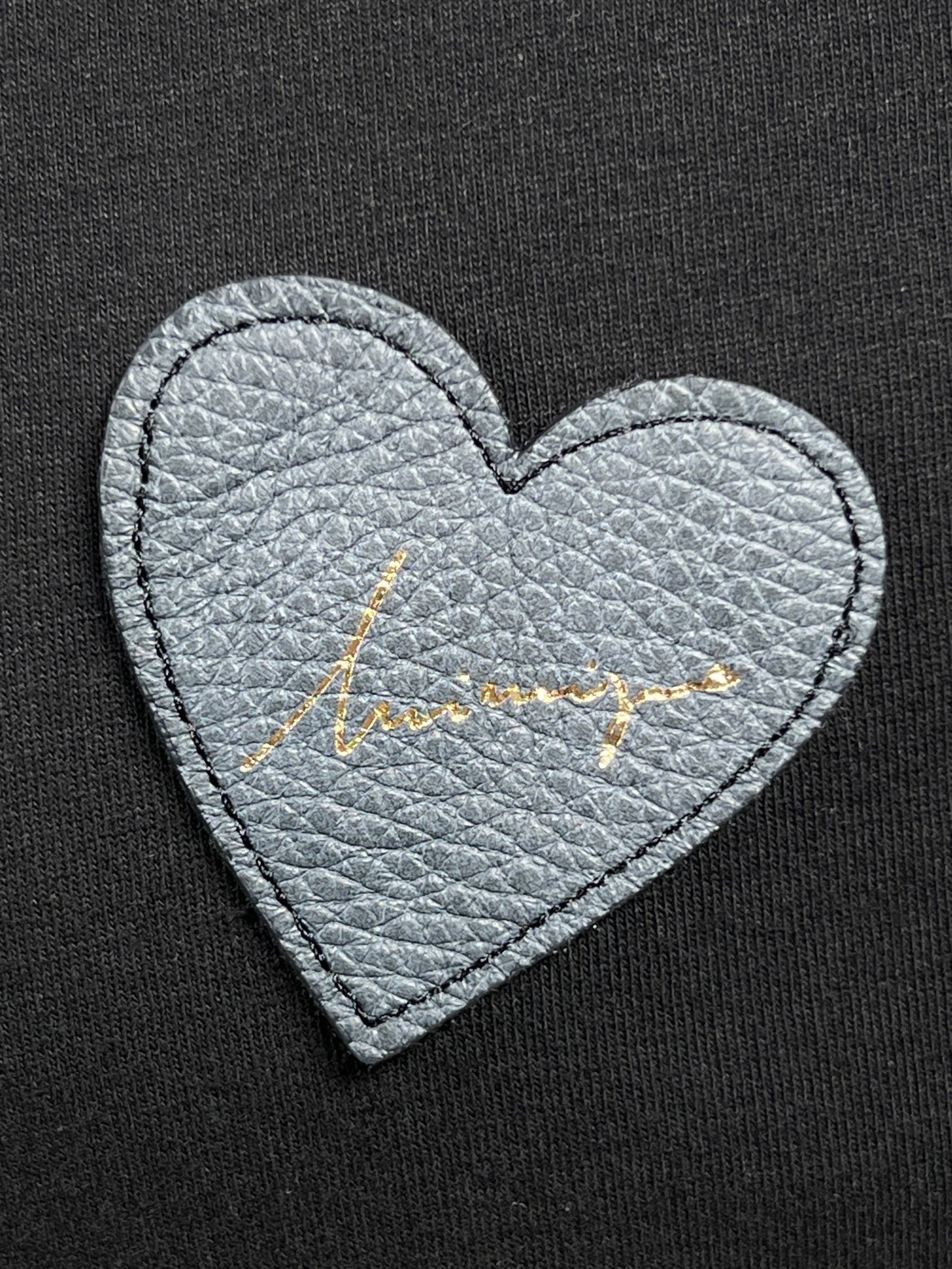 A grey heart-shaped leather detail with gold cursive writing adorns the black fabric of this INIMIGO ITS3508 HEART PATCH T-SHIRT BLACK, which is made from 100% cotton.
