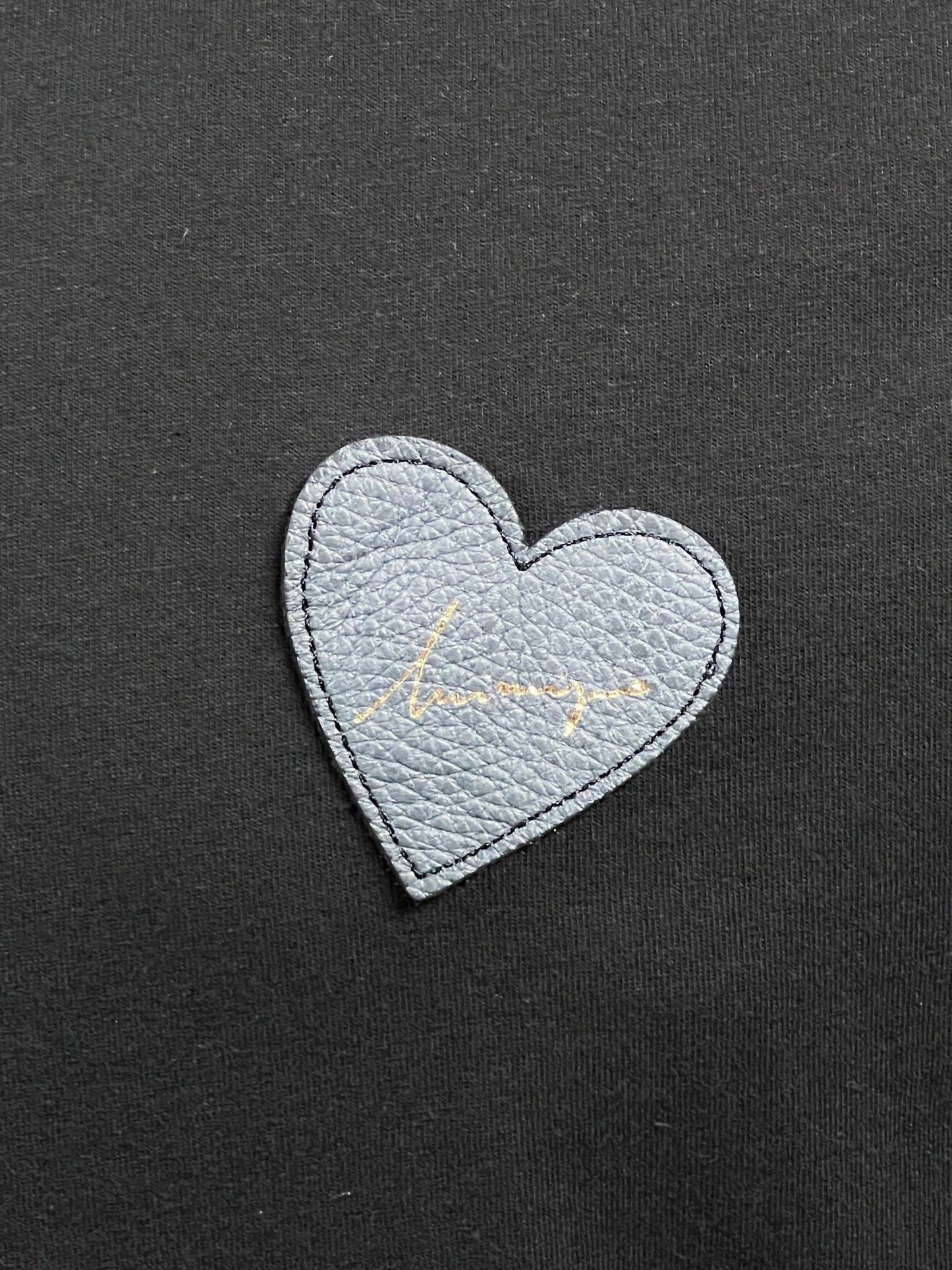 A gray heart-shaped patch with a signature in gold is sewn onto 100% cotton black fabric, adding a touch of elegance to the INIMIGO ITS3508 HEART PATCH T-SHIRT BLACK.