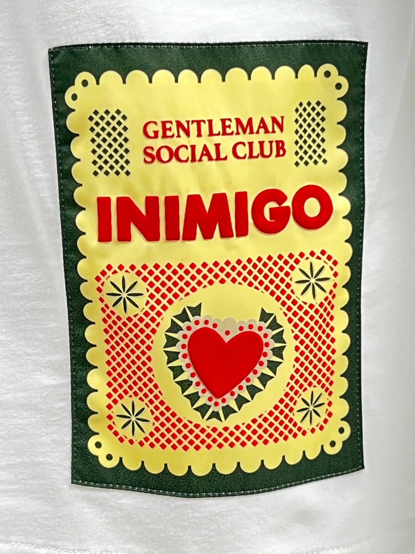 Close-up of the design on a white INIMIGO ITS3539 MEXICAN T-SHIRT WHITE. The 100% cotton fabric showcases "GENTLEMAN SOCIAL CLUB INIMIGO" in red and yellow, with red and green decorative elements and a heart in the center.