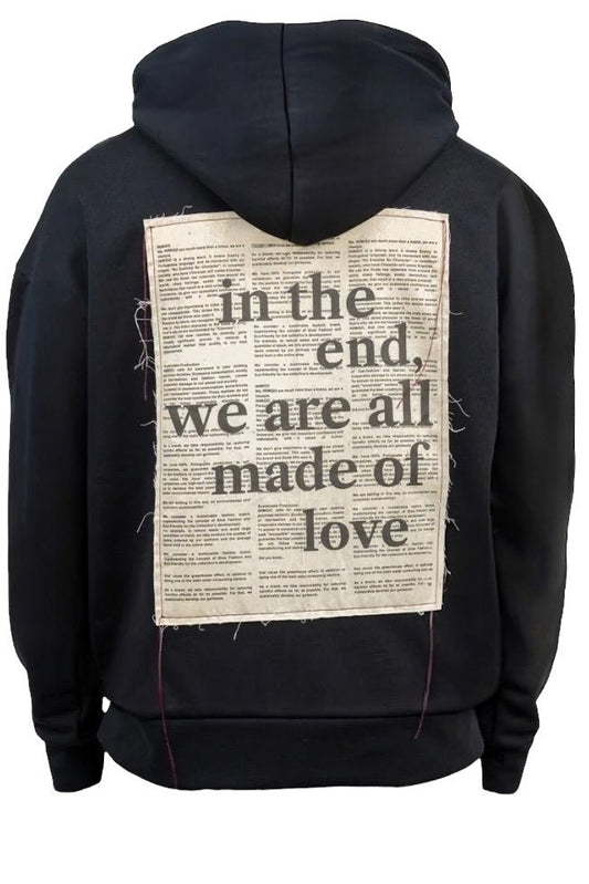 The Inimigo Letter Oversized Hoodie Dk Black by INIMIGO is an oversized black hoodie crafted from organic cotton, featuring the text "In the end, we are all made of love" on the back against a newspaper-style background.