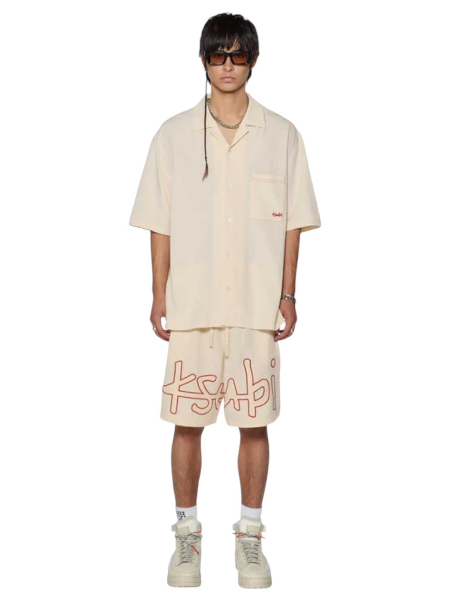 A person stands against a plain white background, wearing Ksubi 1999 Diner ss Shirt Ecru sunglasses and oversized shorts branded with KSUBI, paired with matching socks and white sneakers.