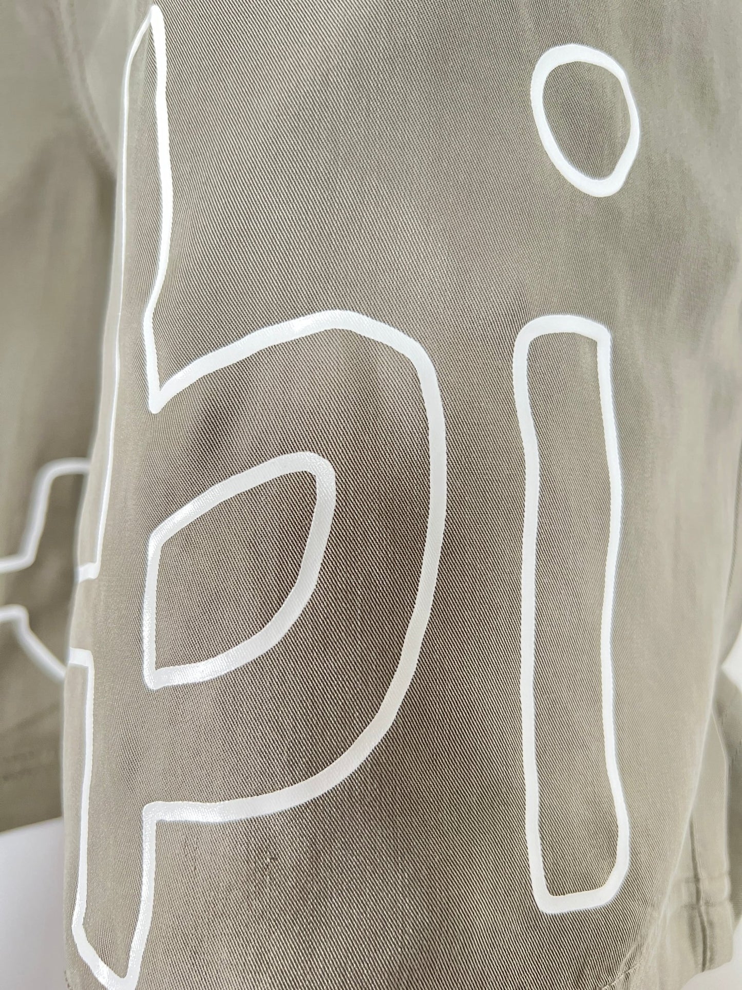 Close-up of a khaki tencel fabric with large white characters printed on it. The visible characters are "p" and "i" on light brown or beige material, reminiscent of the stylish simplicity found in KSUBI 1999 LIFE SHORT OUTBACK GREEN by KSUBI.