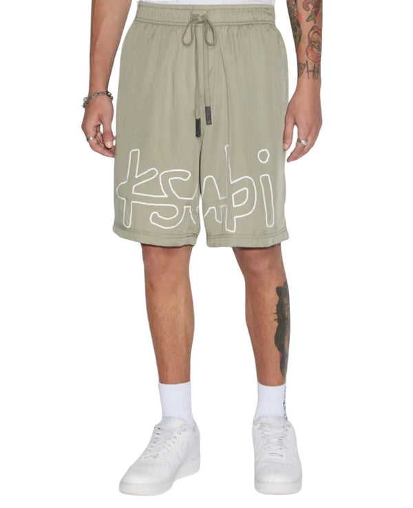 Man wearing KSUBI 1999 LIFE SHORT OUTBACK GREEN with the white "KSUBI" logo, white socks, and white sneakers. Right arm and left calf tattoos are visible.