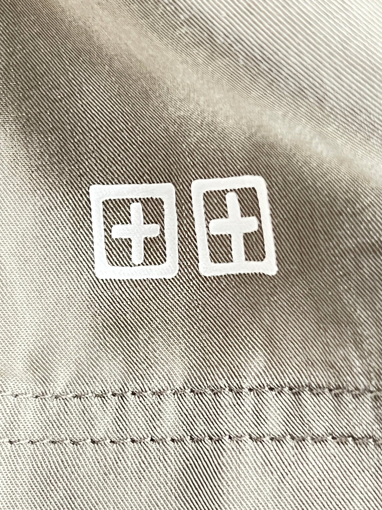 Close-up of two white cross symbols within squares, printed on a textured khaki tencel fabric surface with stitching visible on the KSUBI 1999 LIFE SHORT OUTBACK GREEN by KSUBI.