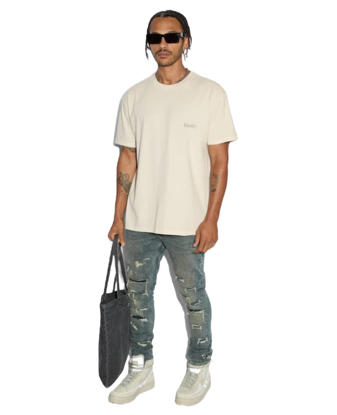 A man in an oversized KSUBI ALL DAY BIGGIE SS TEE JET DESERT TAN and distressed jeans holds a black tote bag. He wears sunglasses and white sneakers, exuding an edgy style.