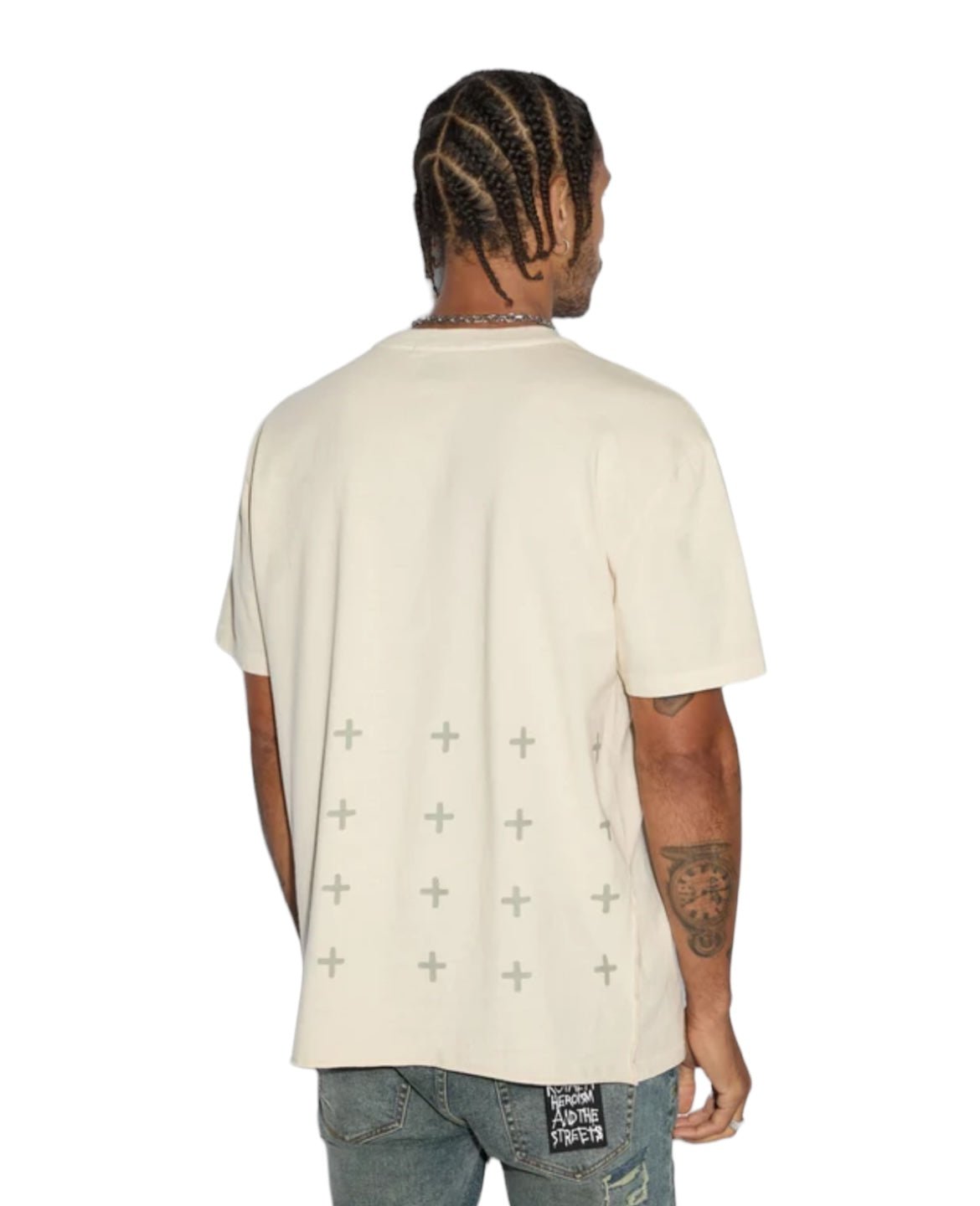 A person with braided hair is wearing an oversized, light-colored KSUBI ALL DAY BIGGIE SS TEE JET DESERT TAN from KSUBI featuring a pattern of crosses and blue jeans with a patched design on the back pocket. The person, exuding an edgy style, is facing away from the camera.