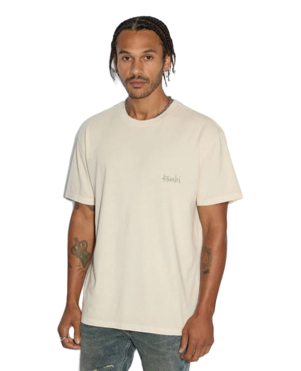 A man with braided hair wearing an oversized KSUBI ALL DAY BIGGIE SS TEE JET DESERT TAN and blue jeans stands with a neutral expression. The T-shirt has the word "KSUBI" on it. He has tattoos on his right arm, adding to his edgy style.