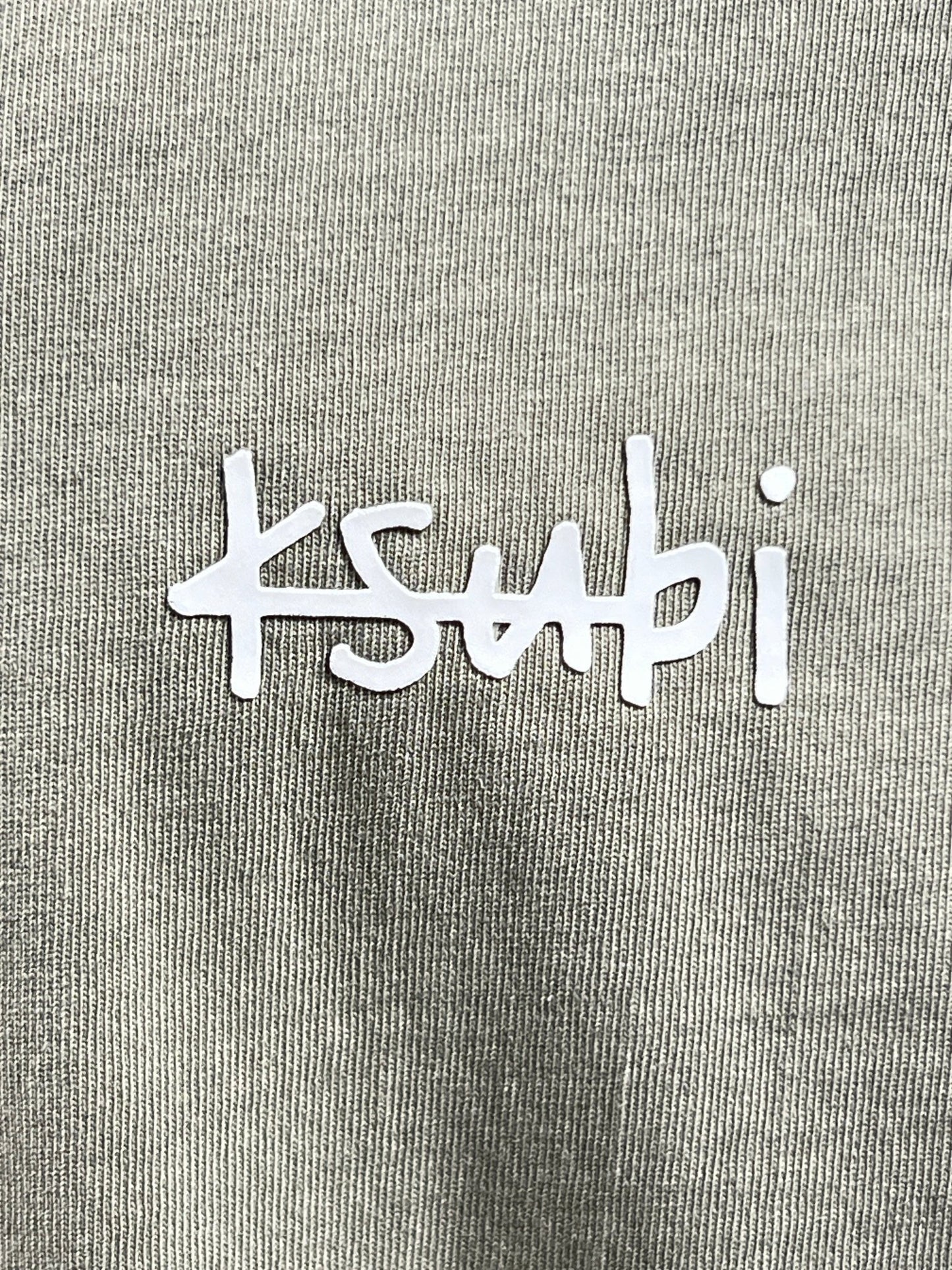 Close-up of a gray premium cotton jersey t-shirt, featuring white "KSUBI ALL DAY BIGGIE TEE OUTBACK GRN" text logo.