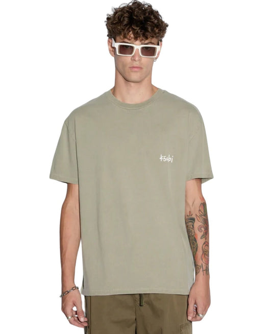 A man wearing white sunglasses, an oversized KSUBI ALL DAY BIGGIE TEE OUTBACK GRN with a Ksubi logo on the left chest, and khaki pants. He has a colorful tattoo on his left arm and is standing against a white background.