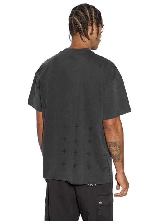 A person with braided hair is standing with their back to the camera, wearing an oversized dark gray KSUBI ANGELIK EKCESS SS TEE ACID BLACK made of heavyweight cotton jersey featuring cross patterns on the back, paired with black pants.