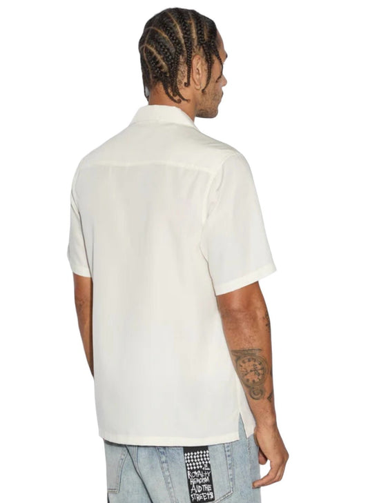 A person with braided hair is seen from behind, wearing the Ksubi Angelik Resort Shirt in white along with denim jeans that feature an angel artwork print patch on the back pocket.