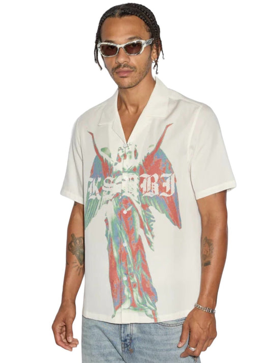 A person wearing the Ksubi Angelik Resort Shirt in white, characterized by a colorful graphic design with angel artwork print from the brand KSUBI, stands against a plain background.