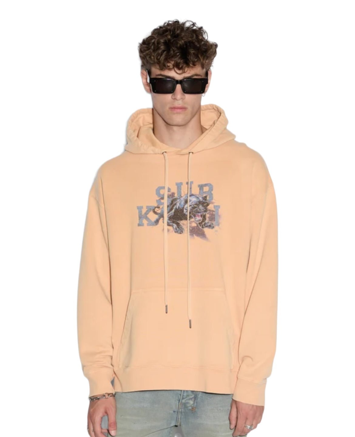 Person wearing a streetwear-inspired KSUBI APEX BIGGIE HOODIE FLARE ORANGE, blue jeans, dark sunglasses, and a bracelet.