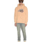A person stands wearing a sun-faded flare orange KSUBI APEX BIGGIE HOODIE with "Club Kava" printed on it, light blue jeans, white high-top shoes, and dark sunglasses—an effortless streetwear look.