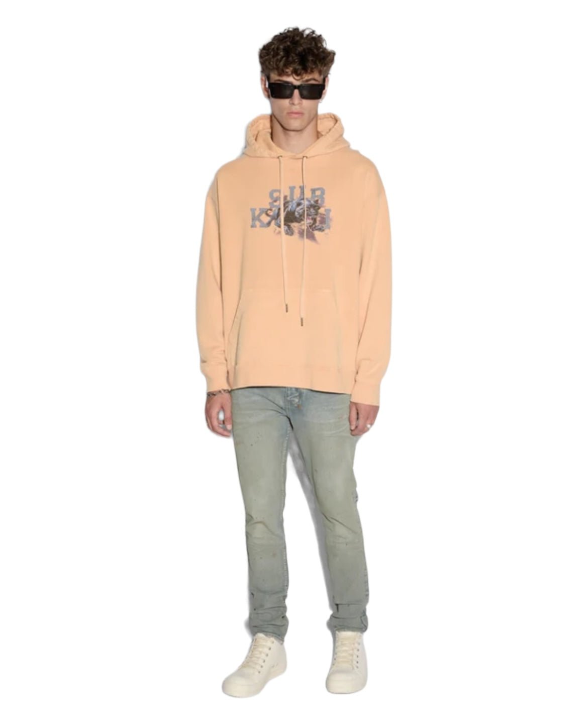 A person stands wearing a sun-faded flare orange KSUBI APEX BIGGIE HOODIE with "Club Kava" printed on it, light blue jeans, white high-top shoes, and dark sunglasses—an effortless streetwear look.