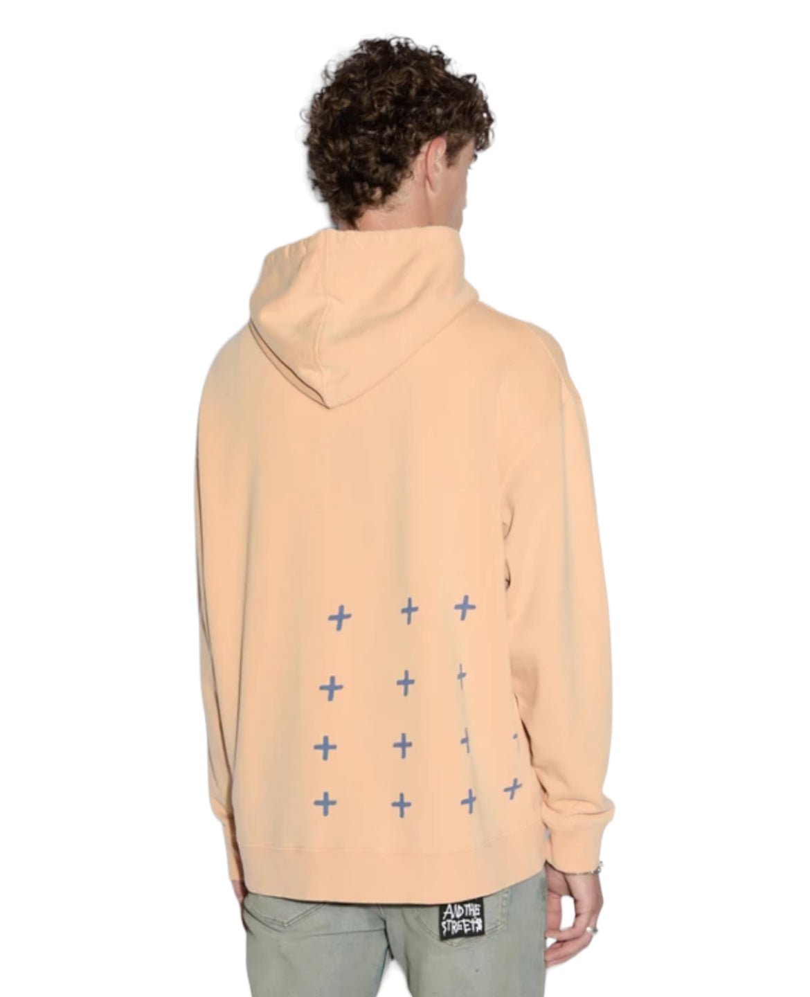 A person with curly hair, facing away from the camera, wears an oversized KSUBI APEX BIGGIE HOODIE FLARE ORANGE adorned with small blue plus signs on the back and light gray pants, epitomizing relaxed streetwear style.