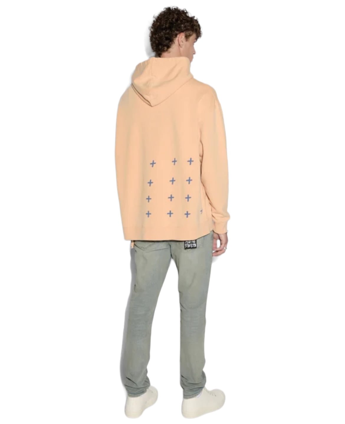 A person wearing a sun-faded KSUBI APEX BIGGIE HOODIE FLARE ORANGE with a pattern of plus signs on the back, grey pants, and white shoes, standing and facing away. This streetwear look is both casual and effortlessly stylish.