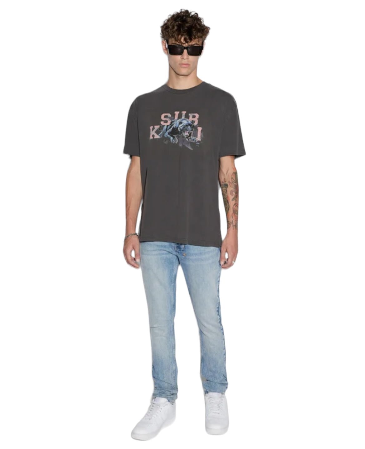 Dressed in a Ksubi Apex Biggie Ss Tee Faded Black from KSUBI, paired with light blue jeans, white shoes, and sunglasses, a person embodies street style against a white background.