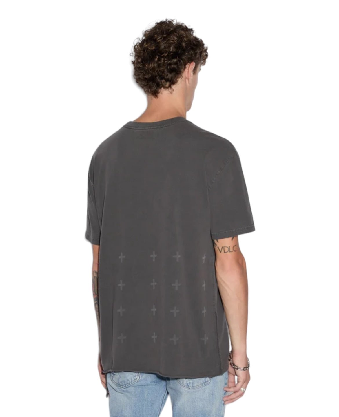 A person with curly hair is shown from the back, wearing a KSUBI APEX BIGGIE SS TEE FADED BLACK, an oversized fit T-shirt by KSUBI with a pattern of small crosses on the lower back, and light blue jeans, embodying an effortless street style.