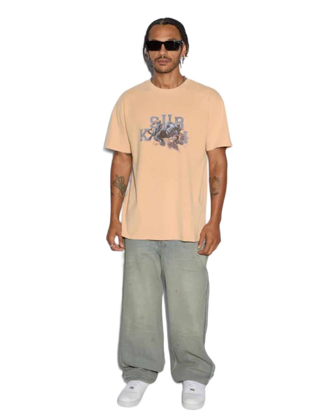 A man confidently stands against a white backdrop in an oversized KSUBI Apex Biggie Ss Tee in Flare Orange, sunglasses, baggy gray pants, and white sneakers.