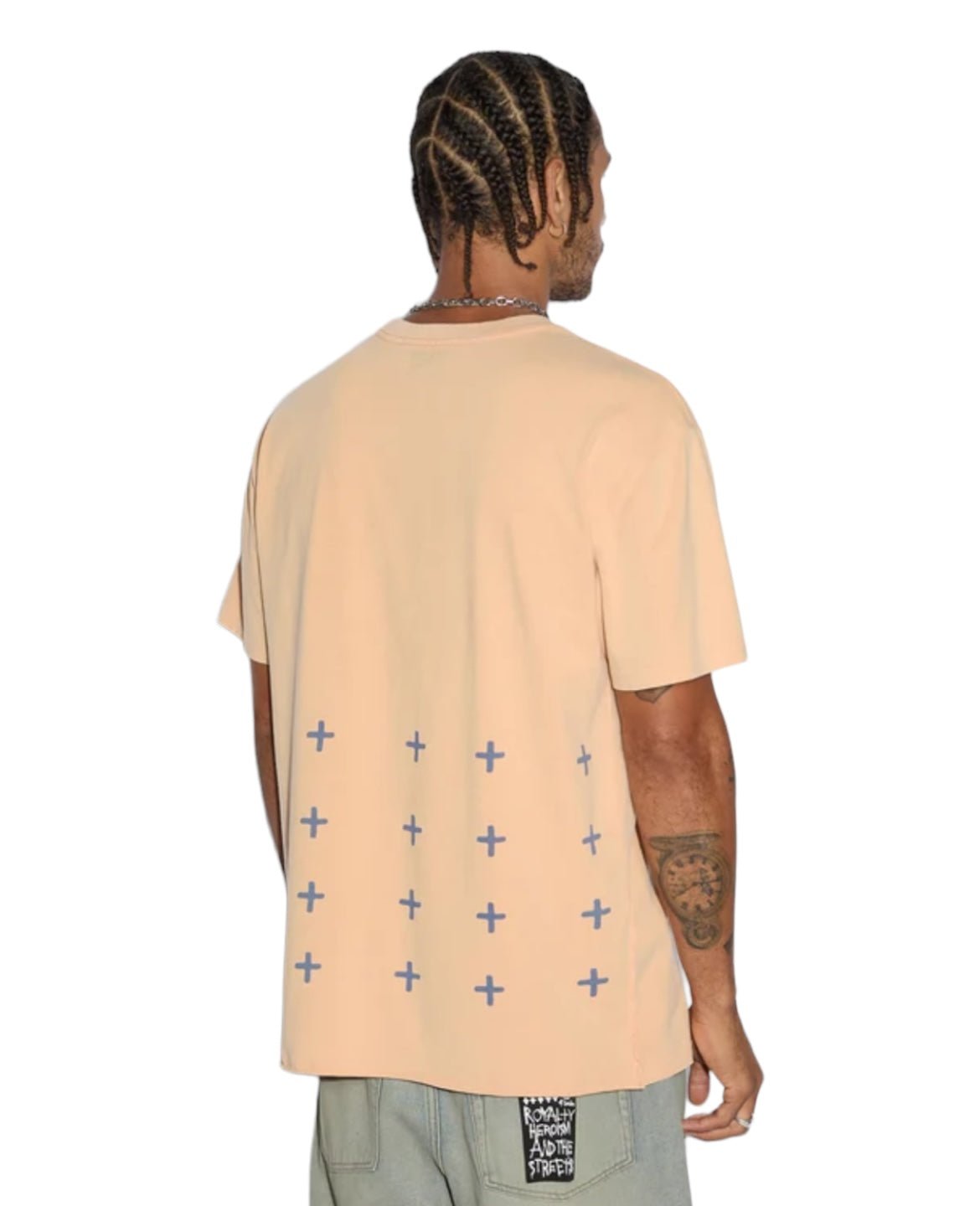 A person with braided hair wears an oversized KSUBI Apex Biggie Ss Tee in sun-faded orange, featuring small blue crosses on the back, paired perfectly with light-colored shorts.