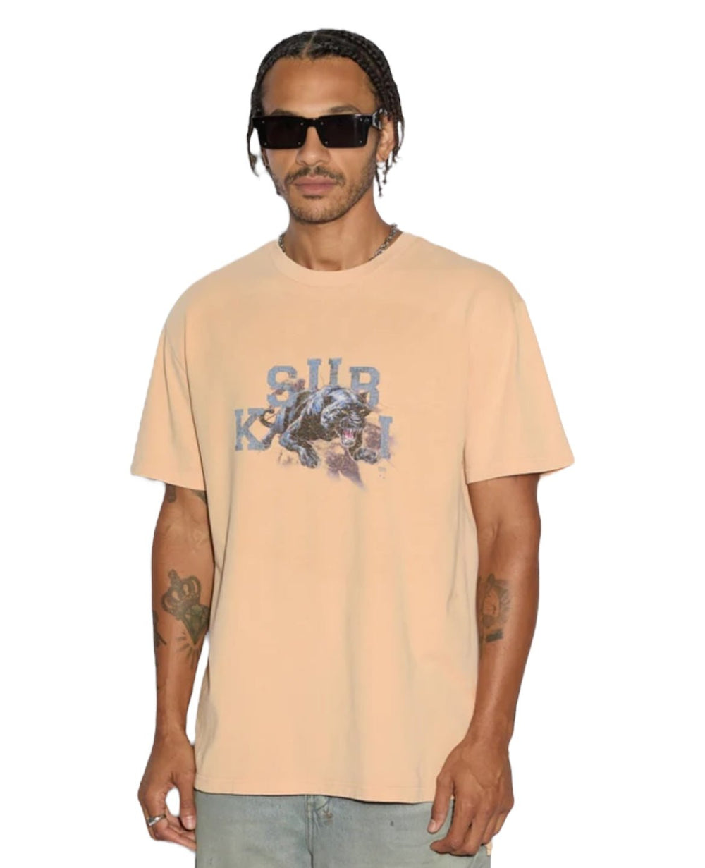 A person in sunglasses and light blue jeans wears a sun-faded KSUBI Apex Biggie SS Tee Flare Orange, featuring a graphic design, against a white background.