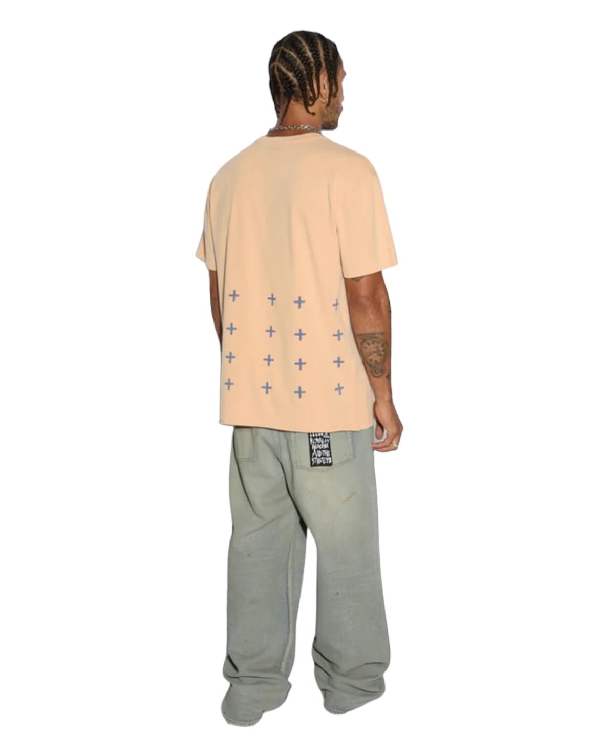 A man with braided hair wearing an oversized KSUBI APEX BIGGIE SS TEE FLARE ORANGE made from premium cotton jersey, featuring small cross patterns on the back, and gray pants stands with his back to the camera.