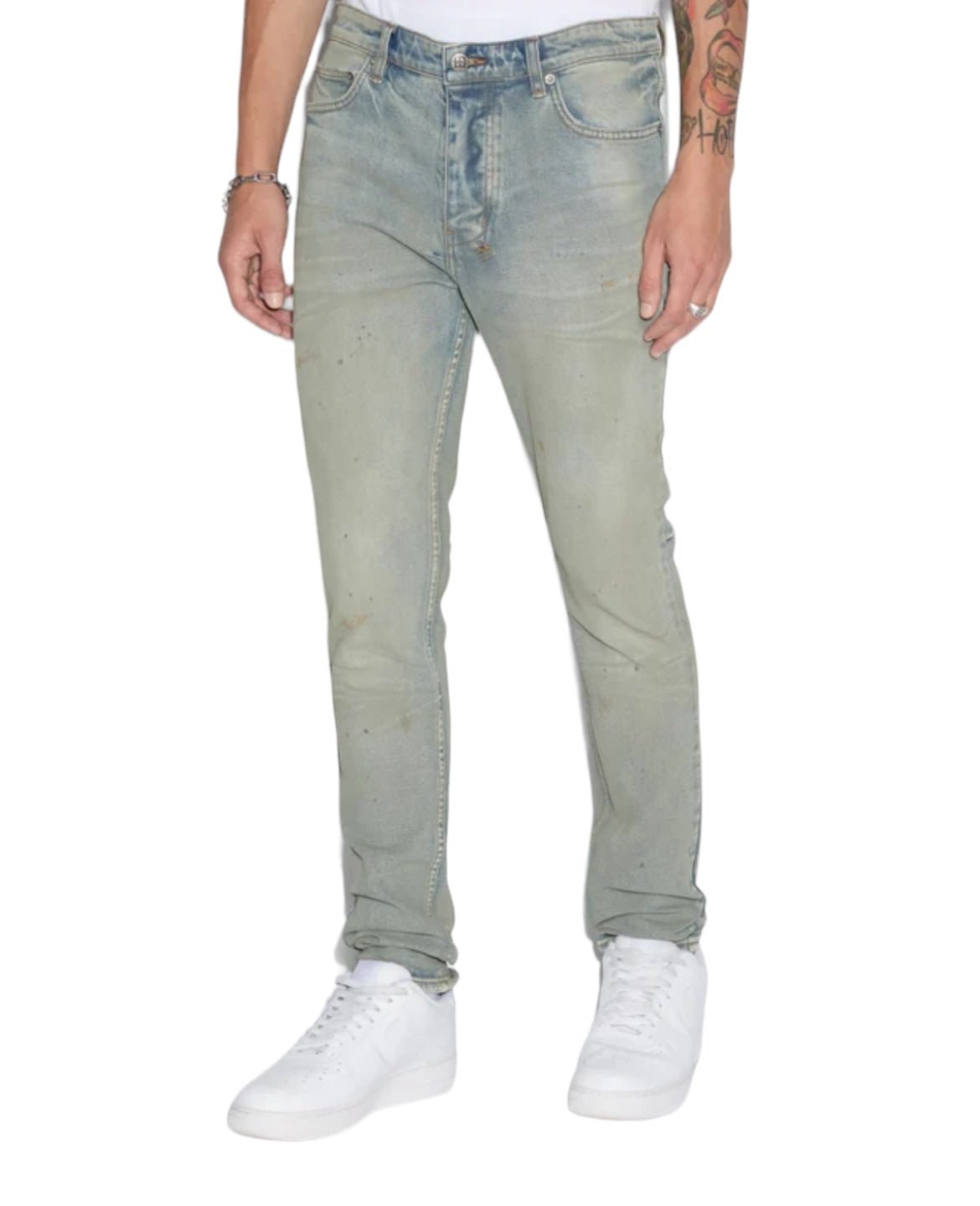 Person wearing slim tapered jeans in light-washed KSUBI CHITCH MECHANIK DENIM from KSUBI, a white shirt tucked in, white sneakers, and a bracelet. Only the torso and legs are visible. One arm has tattoos.