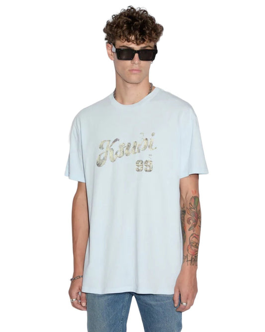 A person with curly hair wearing sunglasses, a light blue KSUBI CLUBHOUSE BIGGIE TEE SHALLOWS BLU with "Ksubi 99" in faded print, and jeans. They have tattoos on their right forearm.