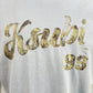 A close-up of a white KSUBI CLUBHOUSE BIGGIE TEE SHALLOWS BLU with the number '99' printed in a distressed, gold-colored script on premium cotton jersey.