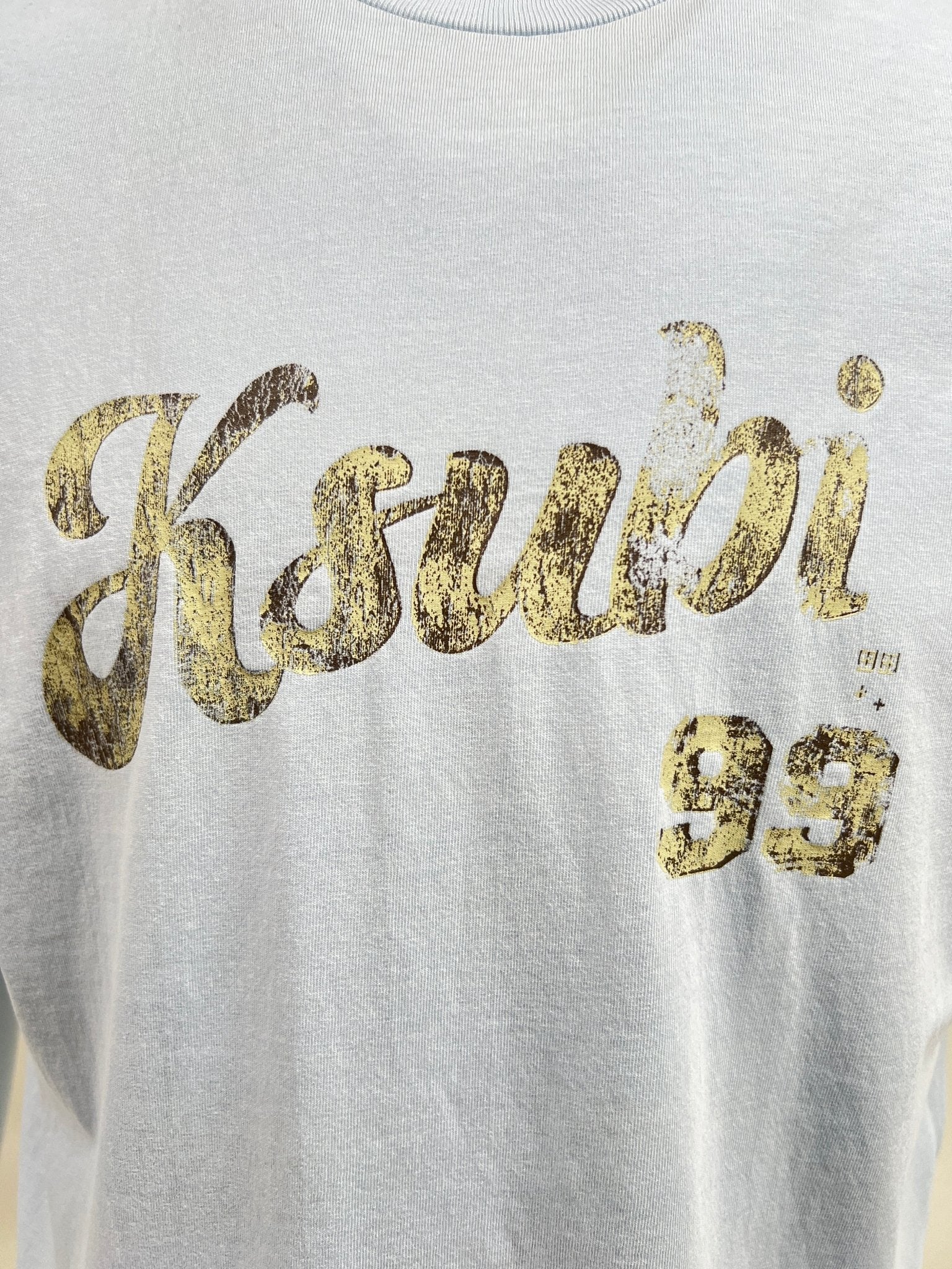 A close-up of a white KSUBI CLUBHOUSE BIGGIE TEE SHALLOWS BLU with the number '99' printed in a distressed, gold-colored script on premium cotton jersey.