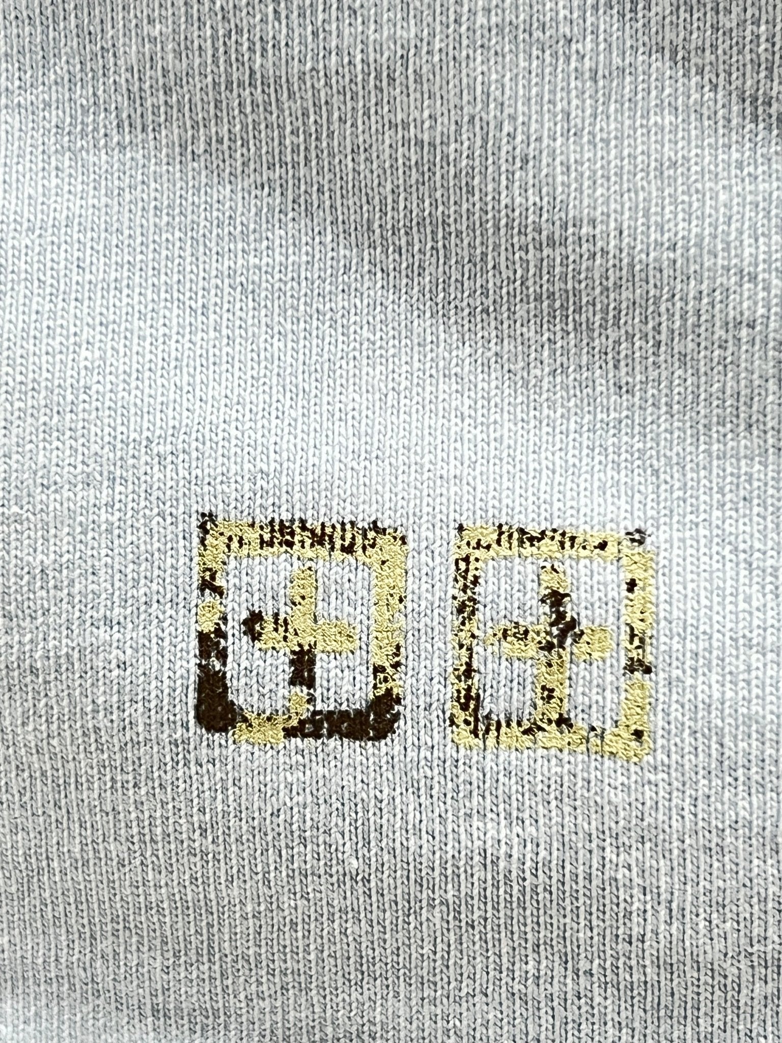 Close-up of a gray fabric from the KSUBI CLUBHOUSE BIGGIE TEE SHALLOWS BLU by KSUBI, showcasing premium cotton jersey with two square symbols featuring internal cross-like designs in yellow and black.