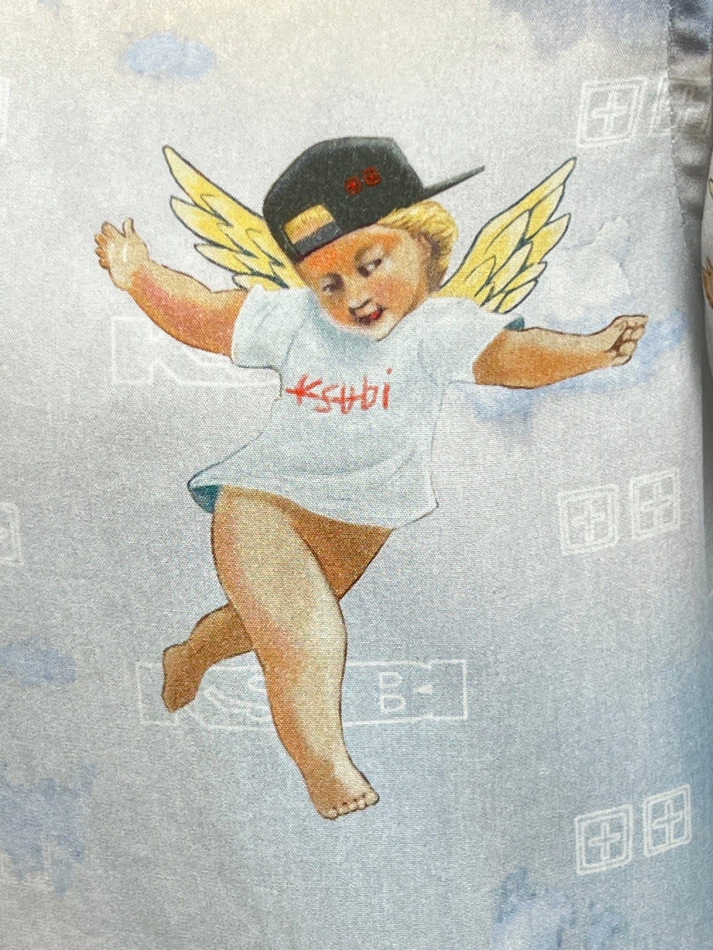 Illustration of a playful cherub in a KSUBI Dreamers Resorts Shirt Multi and black cap, set against a dreamy, cloudy backdrop. Yellow wings add to the whimsical charm.