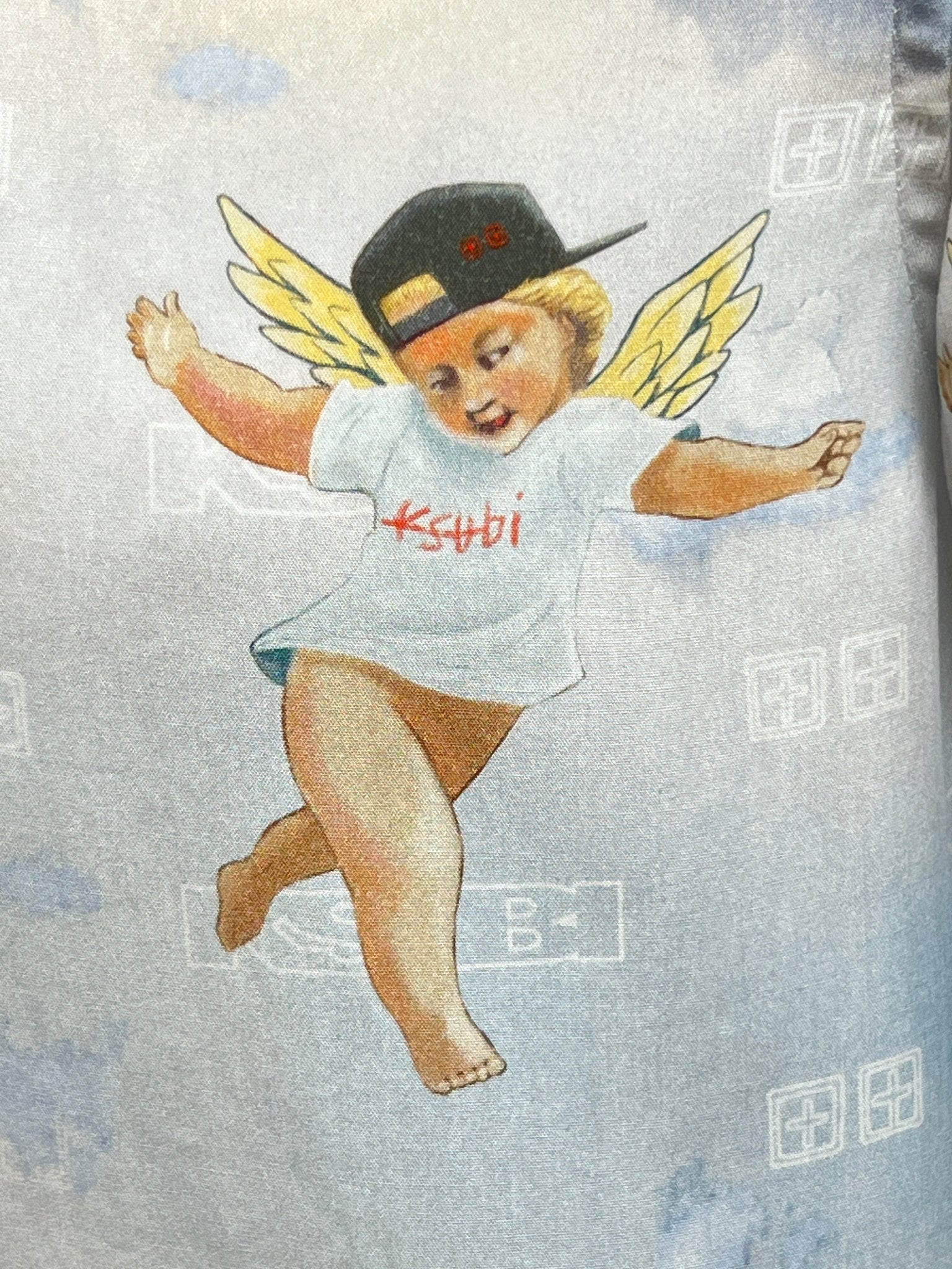 Illustration of a playful cherub in a KSUBI Dreamers Resorts Shirt Multi and black cap, set against a dreamy, cloudy backdrop. Yellow wings add to the whimsical charm.