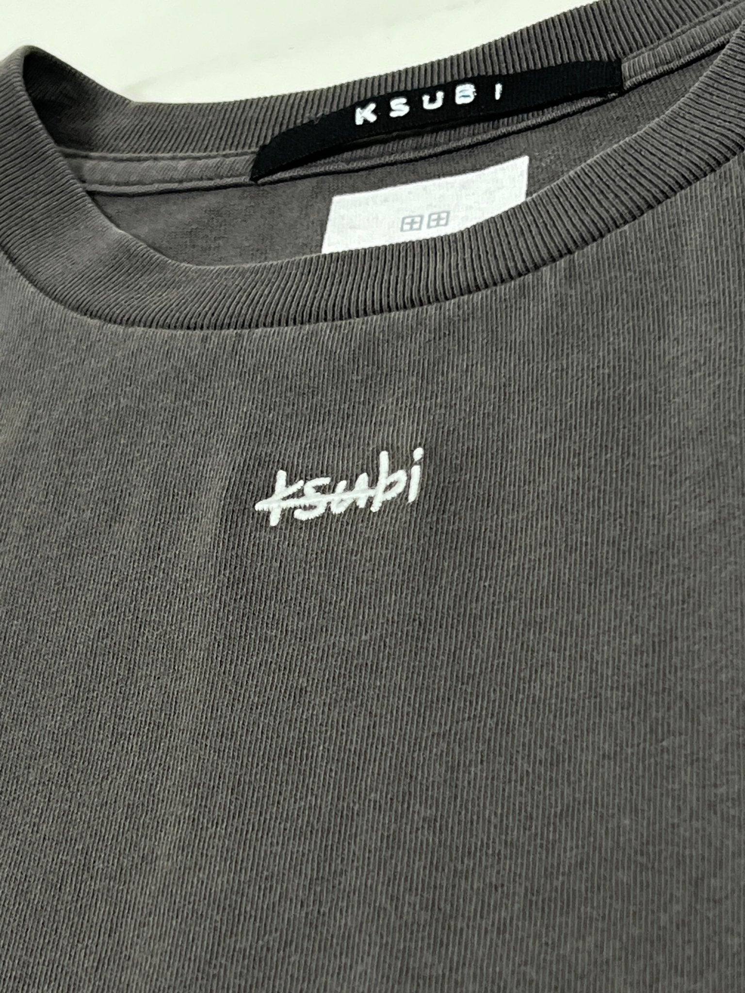 Close-up of a gray Ksubi sweatshirt, showcasing the neck label and embroidered chest logo, made from 100% cotton and echoing the understated style of the Ksubi Eagle Biggie SS Tee in faded black.