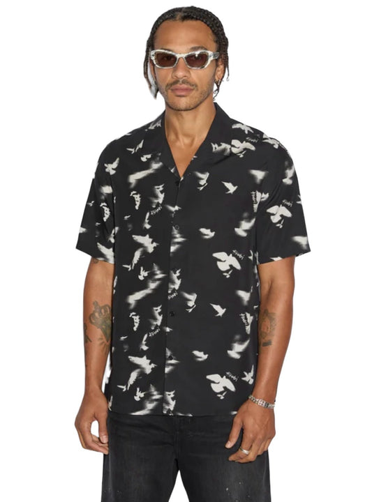 A person with braided hair, wearing sunglasses and the Ksubi Flight Resort SS Shirt in premium tencel fabric, stands facing the camera. The black short-sleeve shirt, featuring a white bird pattern and Ksubi branding, is paired perfectly with dark pants.