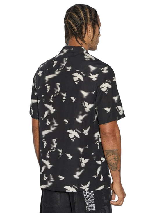 A man with braids wearing the Ksubi Flight Resort SS Shirt in black, featuring white bird prints, is viewed from the back. The premium Tencel fabric of the shirt pairs seamlessly with his black pants, which are adorned with subtle Ksubi branding near the hem.