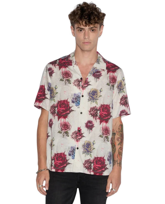 A man with curly hair and tattoos on his left arm is wearing a KSUBI KSUBI GRAFF ROSE RESORT SHIRT MULTI made of premium tencel, featuring a Ksubi rose print in red and blue, paired with black pants.