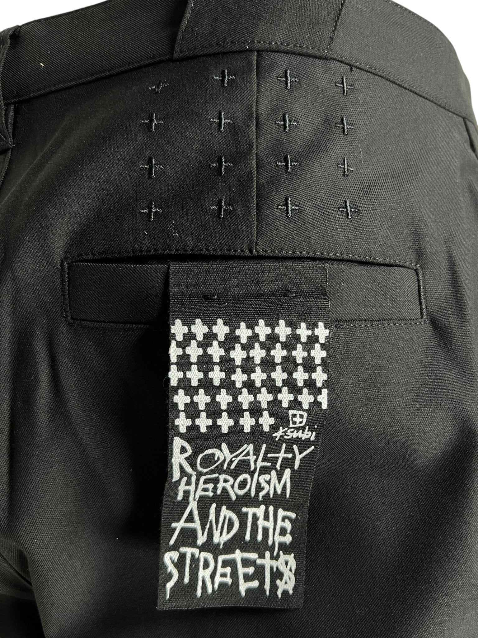 Back view of Ksubi Melrose Suit Pant Black featuring cross stitching and a patch that reads "ROYALTY HEROISM AND THE STREETS," made by KSUBI from a premium polyester, viscose, and elastane blend for comfort and style.