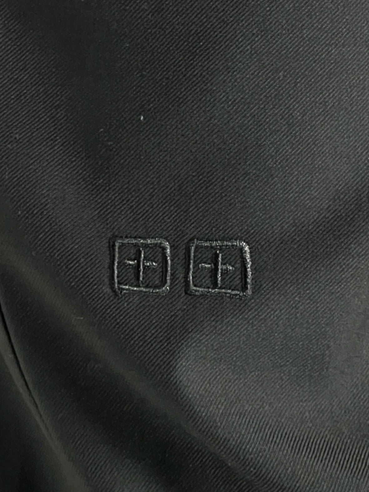 A close-up of the black Ksubi Melrose Suit Pant by KSUBI displays two small embossed square symbols with stylized cross designs.