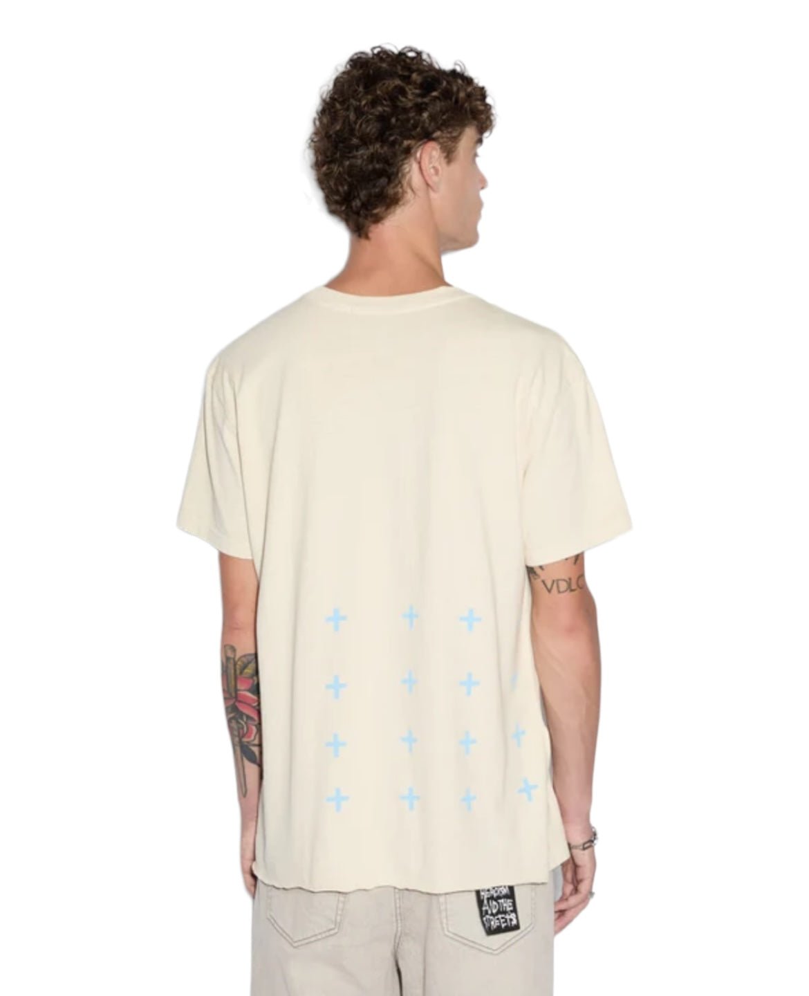 A person with curly hair is seen from the back, wearing a KSUBI PARADISE BIGGIE TEE DUSTY DESERT TAN adorned with blue crosses, paired with tan beige pants. They have tattoos on their right arm and a patch on their back pocket.